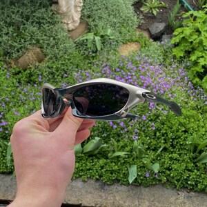 May include: Silver and black sunglasses with dark lenses. The sunglasses have a sporty design with a curved frame and arms. The lenses are dark, providing protection from the sun. The sunglasses are held up against a background of green foliage and purple flowers.