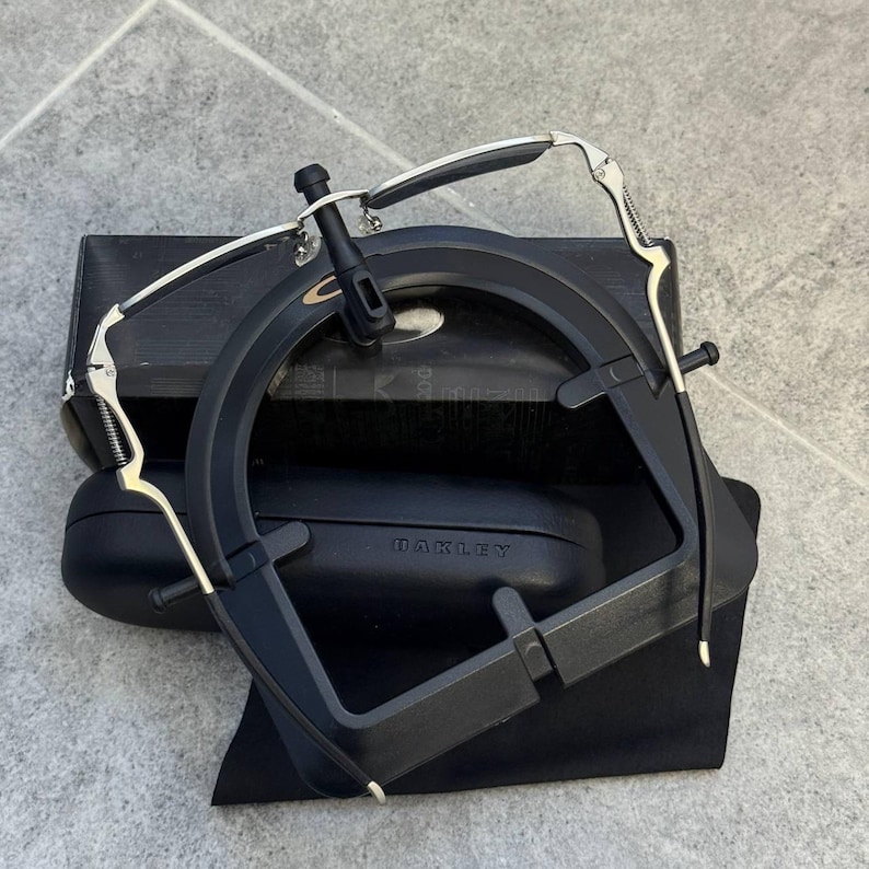May include: Black and silver Oakley sunglasses with a black case and a black cloth bag. The sunglasses have a unique frame design with silver accents and a black bridge. The case has the brand name "OAKLEY" printed on it.