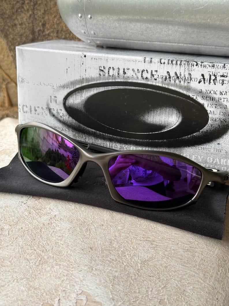 May include: Silver-framed sunglasses with purple lenses rest on a black cloth. The lenses reflect a purple hue. A silver box with the text "SCIENCE AND ART" is in the background.