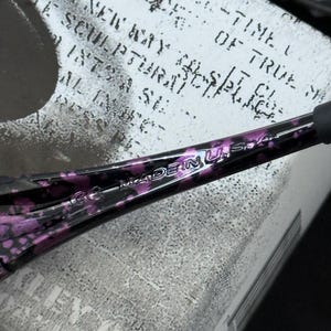 Puede incluir: Primer plano de una patilla de gafas con estampado negro y morado. El texto "MADE IN U.S.A." es visible. La patilla descansa sobre una superficie blanca con texto negro descolorido.