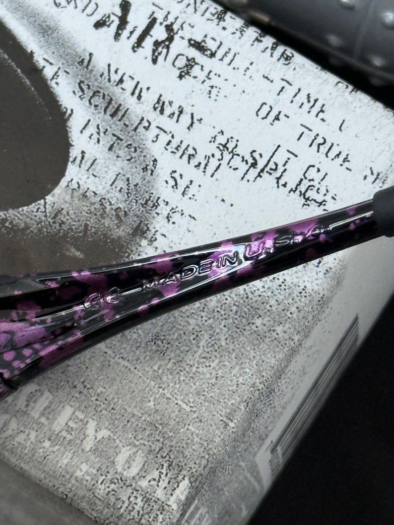 Puede incluir: Primer plano de una patilla de gafas con estampado negro y morado. El texto "MADE IN U.S.A." es visible. La patilla descansa sobre una superficie blanca con texto negro descolorido.