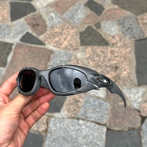 May include: Gray sunglasses with dark lenses. The sunglasses have a unique, angular design with a curved temple arm. The lenses reflect the surrounding environment. The frame is a matte gray color.