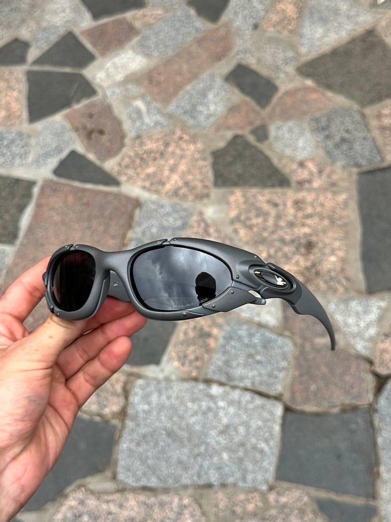 May include: Gray sunglasses with dark lenses. The sunglasses have a unique, angular design with a curved temple arm. The lenses reflect the surrounding environment. The frame is a matte gray color.