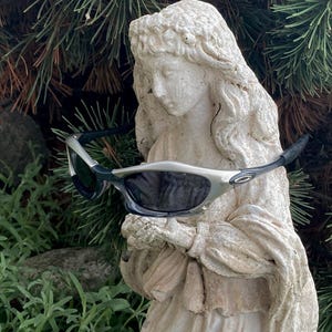 May include: Silver and black sunglasses with dark lenses rest on a white statue of a woman. The sunglasses have a sporty design with a silver frame and black arms. The statue is weathered and stands in front of green foliage.