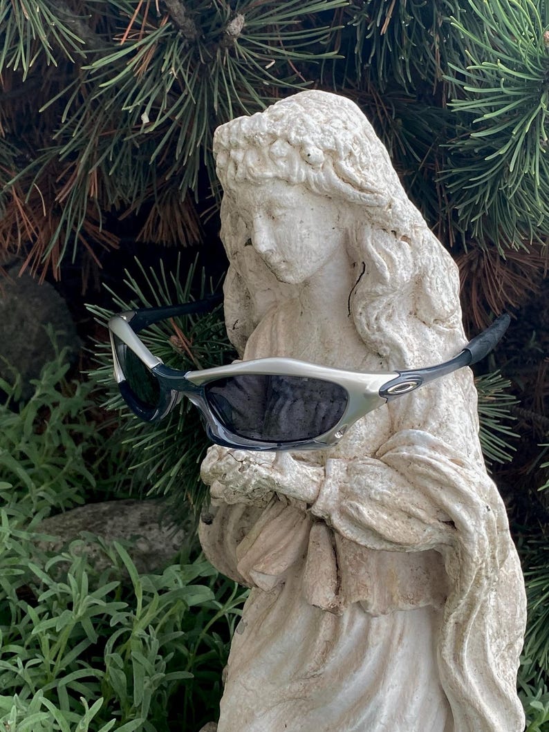 May include: Silver and black sunglasses with dark lenses rest on a white statue of a woman. The sunglasses have a sporty design with a silver frame and black arms. The statue is weathered and stands in front of green foliage.