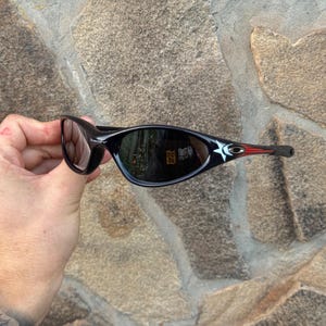 May include: Black sunglasses with a sleek, curved design. The frames are black with a red accent on the temples. The lenses are dark, reflecting the surroundings. The sunglasses are held against a stone wall.
