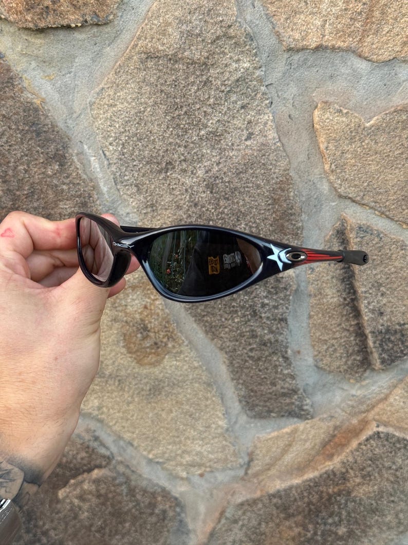 May include: Black sunglasses with a sleek, curved design. The frames are black with a red accent on the temples. The lenses are dark, reflecting the surroundings. The sunglasses are held against a stone wall.