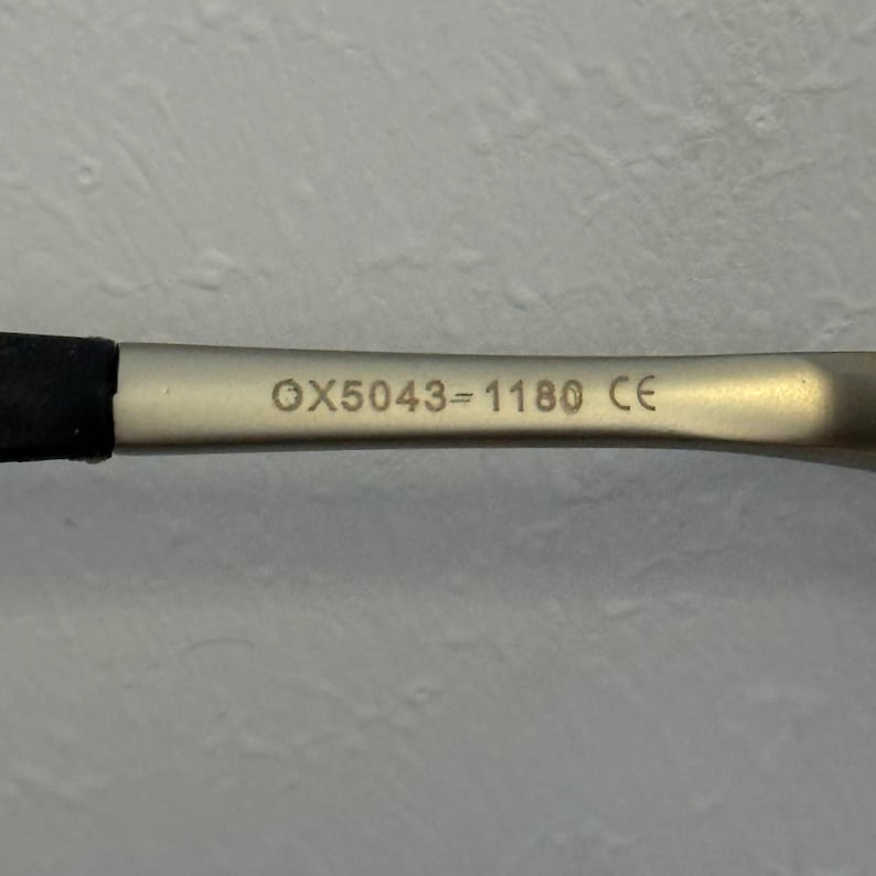 May include: A close-up shot of a silver metal object, featuring the etched text "OX5043-1180 CE". The object has a black, rubber-like section on the left. The background is a plain, off-white wall.
