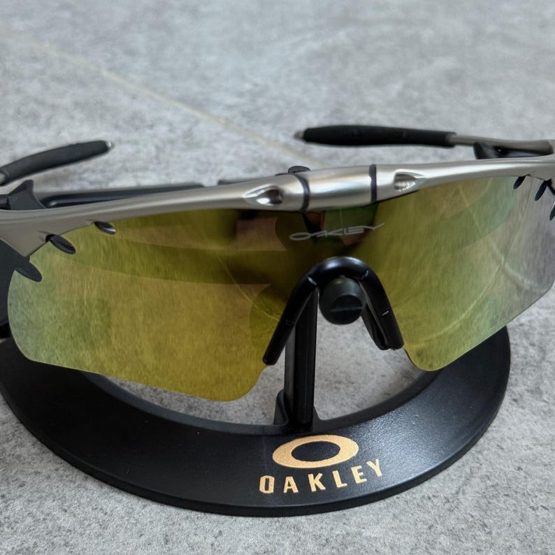 May include: Silver-framed sunglasses with yellow-tinted lenses, displayed on a black stand. The stand features the brand name "OAKLEY" in gold lettering. The sunglasses have a sleek, modern design.