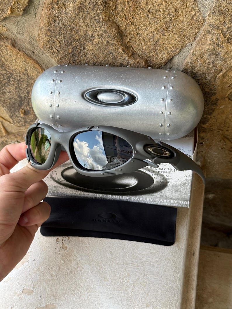 May include: Gray Oakley sunglasses with a matching case and black carrying pouch. The sunglasses have a curved frame and reflective lenses. The case is silver with a riveted design and the Oakley logo. The lenses reflect the sky.