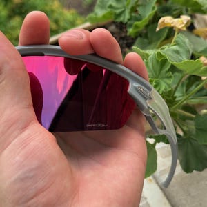 May include: Gray sunglasses with red-tinted lenses are held in a hand. The lenses reflect a pink and purple hue. The frame is a light gray color. The brand name "PRIZM" is visible on the lens.