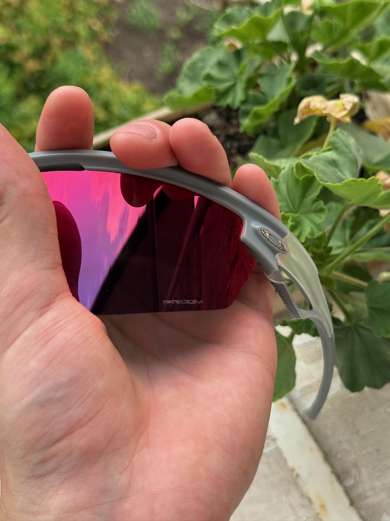 May include: Gray sunglasses with red-tinted lenses are held in a hand. The lenses reflect a pink and purple hue. The frame is a light gray color. The brand name "PRIZM" is visible on the lens.