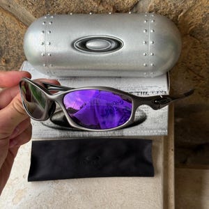 May include: Silver-framed sunglasses with purple mirrored lenses, a black microfiber pouch, and a silver case. The case has a rounded shape with rivets and the brand logo. The lenses reflect a purple hue.