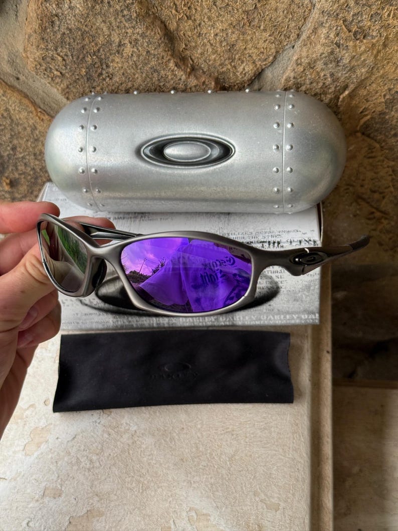 May include: Silver-framed sunglasses with purple mirrored lenses, a black microfiber pouch, and a silver case. The case has a rounded shape with rivets and the brand logo. The lenses reflect a purple hue.