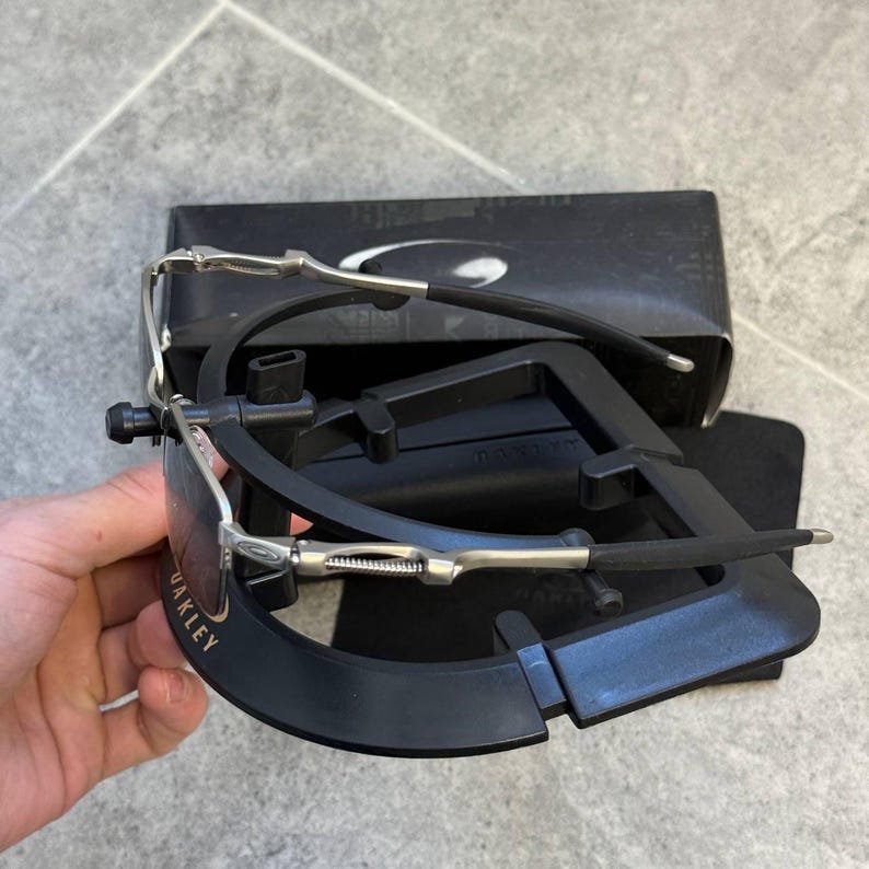 May include: Black Oakley sunglasses with silver detailing and a black case. The sunglasses feature a curved design with gold lettering. The case is black and rectangular.