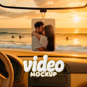 May include: A rectangular car air freshener with a photo of a couple embracing, hanging from a black bar. The background shows a beach scene with a sunset. The words "video MOCKUP" are displayed in white, 3D letters.