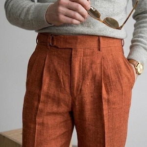 May include: High-waisted trousers in a vibrant burnt orange hue, featuring a textured fabric finish, double front pleats, and a side-adjuster buckle strap at the waistband. The trousers are paired with a light grey long-sleeved sweater.