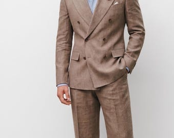 May include: A man wearing a light brown double-breasted suit, light blue shirt, and a pink flower in the lapel. The suit is paired with black dress shoes. The trousers are wide-legged.