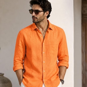 Könnte beinhalten: Ein orangefarbenes Leinenhemd mit Knopfleiste und Brusttasche. Das Hemd wird mit Sonnenbrille und Uhr getragen. Die Ärmel sind hochgekrempelt. Das Hemd wird mit einer weißen Hose kombiniert.