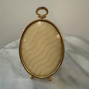 May include: An oval-shaped gold-toned metal picture frame with a smooth, polished finish. The frame features a decorative circular loop at the top and rests on two small, rounded feet. Inside is a cream-colored insert with a wavy pattern.