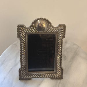 May include: A silver-toned metal picture frame with a rectangular center opening and a rounded top arch. The frame features a raised, repeating diagonal ribbed pattern along the borders, accented by small beaded details and a circular medallion.