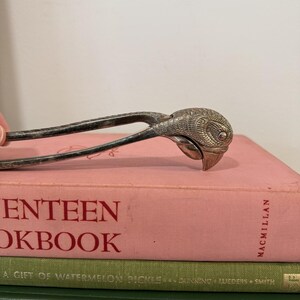 May include: A silver-toned nutcracker with a bird-head design, resting on a stack of cookbooks. The nutcracker's head is intricately detailed, and the books are in shades of pink and green. The top book reads "Seventeen Cookbook".