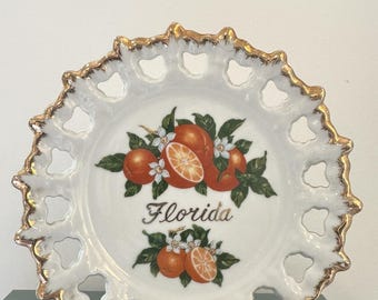 May include: White decorative plate with a gold rim and openwork design. The plate features a colorful illustration of oranges, blossoms, and the word "Florida" in script. A charming souvenir.