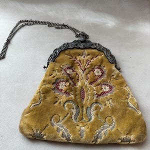 May include: A mustard yellow velvet handbag featuring a woven floral pattern in deep red and grey tones. The bag has an ornate, dark metal frame with a decorative clasp and a thin, matching dark metal chain strap for carrying or wearing.
