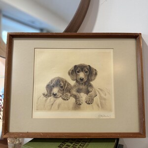 May include: A framed print featuring two dachshund puppies. The sepia-toned artwork depicts the puppies resting on a soft surface. The frame is a warm brown, and the print is matted with a neutral color.