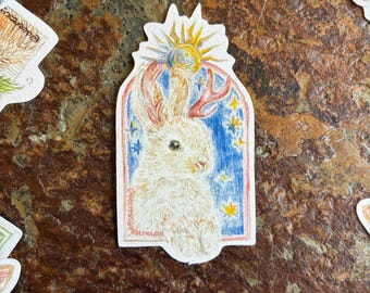 May include: A whimsical sticker featuring a white rabbit with antler-like horns, a sun, and stars against a blue background. The sticker has a decorative border and the artist's signature. Other stickers are visible in the background.