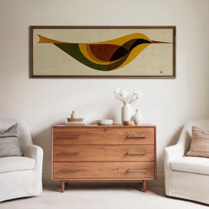Op de afbeelding: Een ingelijst kunstwerk met een gestileerd vogelontwerp in tinten geel, bruin, groen en zwart. Het kunstwerk is geplaatst tegen een neutrale achtergrond en omlijst in donker hout. Daaronder een houten dressoir met drie lades.