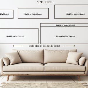 a living room with a large beige sofa, a wooden side table, and a potted plant. The sofa is positioned in front of a wall with a size guide for artwork, providing dimensions for different sizes of framed artworks.