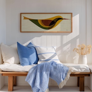 Op de afbeelding: Een houten bank met witte kussens, blauwe kussens en een blauw gestreepte deken. Boven de bank hangt een ingelijst kunstwerk van een gestileerde vogel in geel, bruin en groen. Een witte vaas met gedroogde bloemen staat in de buurt.