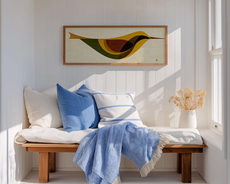 Op de afbeelding: Een houten bank met witte kussens, blauwe kussens en een blauw gestreepte deken. Boven de bank hangt een ingelijst kunstwerk van een gestileerde vogel in geel, bruin en groen. Een witte vaas met gedroogde bloemen staat in de buurt.