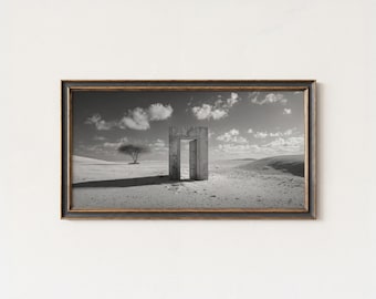 May include: Black and white framed print of a surreal desert landscape. A stone doorway stands open in the center, casting a long shadow. A lone tree is in the distance under a cloudy sky. The frame is dark with gold accents.