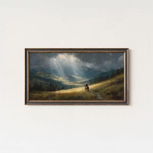 May include: Framed landscape painting depicting a rider on horseback traversing a grassy path. The artwork features a dramatic sky with sun rays illuminating a valley between rolling hills and mountains. The frame is dark with gold accents.