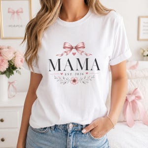 Può includere: T-shirt bianca con la scritta "MAMA" in nero, ornata da un fiocco rosa e un design floreale. Il testo "EST. 2026" è sotto. Un capo d'abbigliamento casual.