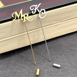 May include: Two lapel pins with initials. One pin is gold-toned with the initials "MR", and the other is silver-toned with the initial "K". The pins have a slender design and are likely used as accessories for jackets or suits.