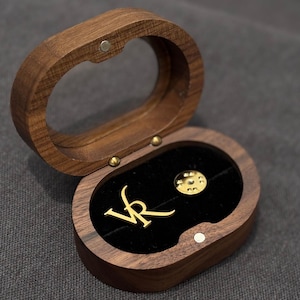 May include: A wooden jewellery box with a hinged lid, open to reveal a black velvet interior. Inside, two gold-coloured pins are displayed: one with the letters "VR" and a round pin. The box is made of dark wood.