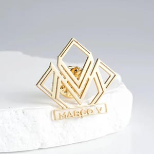 Può includere: Spilla dorata con un design geometrico, composta da forme a diamante e il nome "MARCO V" alla base. La spilla è posta su uno sfondo bianco, evidenziando i suoi dettagli intricati e la lucentezza metallica.