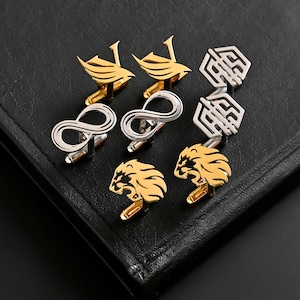 May include: A collection of cufflinks in gold and silver tones, featuring various designs. The cufflinks include bird-like shapes, infinity symbols, geometric patterns, and lion heads. The accessories are displayed on a black leather surface.