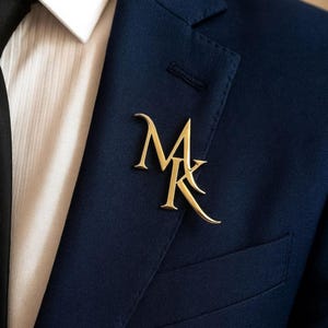 May include: A gold-coloured lapel pin featuring the intertwined initials "MK". The pin is attached to a navy blue suit jacket, worn with a white shirt and a black tie. A sophisticated accessory for formal wear.