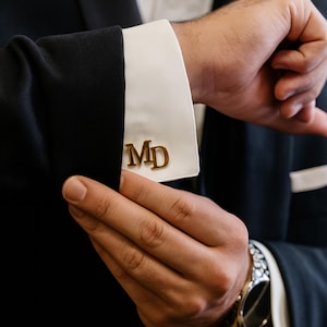 May include: Gold-colored cufflinks with the initials "MD" on a white dress shirt cuff. The cuff is part of a black suit jacket. A watch with a silver band is visible on the wrist.