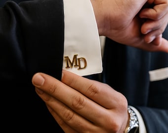 May include: Gold cufflinks bearing the initials "MD" on a white shirt cuff, part of a black suit jacket. A silver wristwatch is visible on the wrist. The image focuses on the details of formal wear.