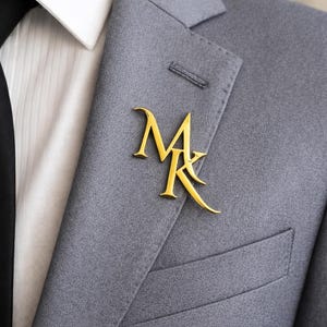 May include: A gold-colored lapel pin in the shape of the letters "MK" is affixed to a gray suit jacket. The pin is positioned on the left lapel, near the pocket. The jacket is worn over a white dress shirt and a black tie.