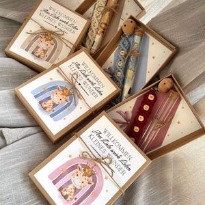 Puede incluir: Cajas de regalo con artículos para bebés. Cada caja contiene una tarjeta decorativa con un diseño de jirafa y arcoíris, y el texto "BIENVENIDA, Del amor, pequeña maravilla." Las cajas también contienen un clip para chupete.