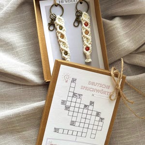Może przedstawiać: Pudełko upominkowe zawierające dwa kremowe breloczki z makramy z mosiężnym osprzętem i kolorowymi koralikami. Breloczki mają napis "IHR WERDET". Dołączona jest również krzyżówka z tekstem "DEUTSCHE SPRICHWÖRTER".