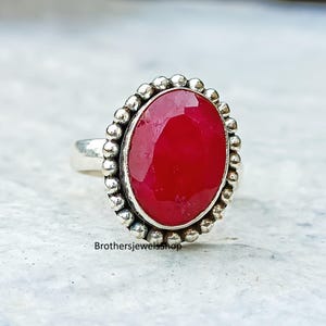 Op de afbeelding: Een zilveren ring met een grote, ovaalvormige, dieprode edelsteen. De edelsteen is omgeven door een kralenrand. De ring heeft een gladde band. De tekst "Brothersjewelsshop" is zichtbaar onderaan de afbeelding.