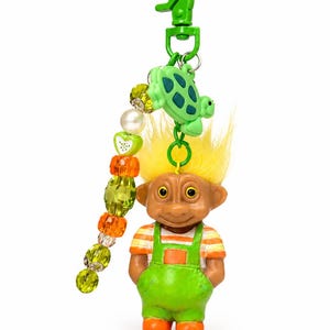 May include: A troll doll keychain featuring bright yellow hair, wearing orange and white striped sleeves under lime green overalls. A green plastic turtle charm and a strand of lime green, orange, and white beads hang from a green plastic clip.