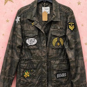 May include: Camouflage-patterned jacket with multiple pockets and decorative patches. Patches include stars, wings, and text that reads "MY ALONE TIME IS FOR YOUR SAFETY" and "NOT MY CIRCUS NOT MY MONKEYS NOT MY PROBLEM". The jacket is hanging on a black hanger.